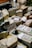 A variety of cheeses are displayed on a market stand, each labeled with pricing. Some of the packages are wrapped in plastic. There are visible price tags, including one for 'Pecorino' and others with varying prices. In the background, there are bottles and other produce items.