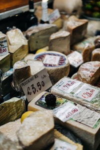 A variety of cheeses are displayed on a market stand, each labeled with pricing. Some of the packages are wrapped in plastic. There are visible price tags, including one for 'Pecorino' and others with varying prices. In the background, there are bottles and other produce items.