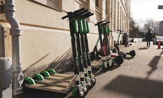 Several electric scooters are lined up against the wall of a building on a sunny day. A person is walking on the sidewalk nearby, with some cars parked along the street. A traffic cone is visible further down the sidewalk.