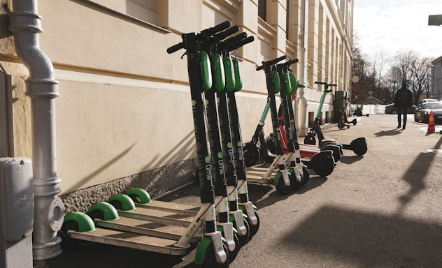 Several electric scooters are lined up against the wall of a building on a sunny day. A person is walking on the sidewalk nearby, with some cars parked along the street. A traffic cone is visible further down the sidewalk.