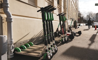 Several electric scooters are lined up against the wall of a building on a sunny day. A person is walking on the sidewalk nearby, with some cars parked along the street. A traffic cone is visible further down the sidewalk.