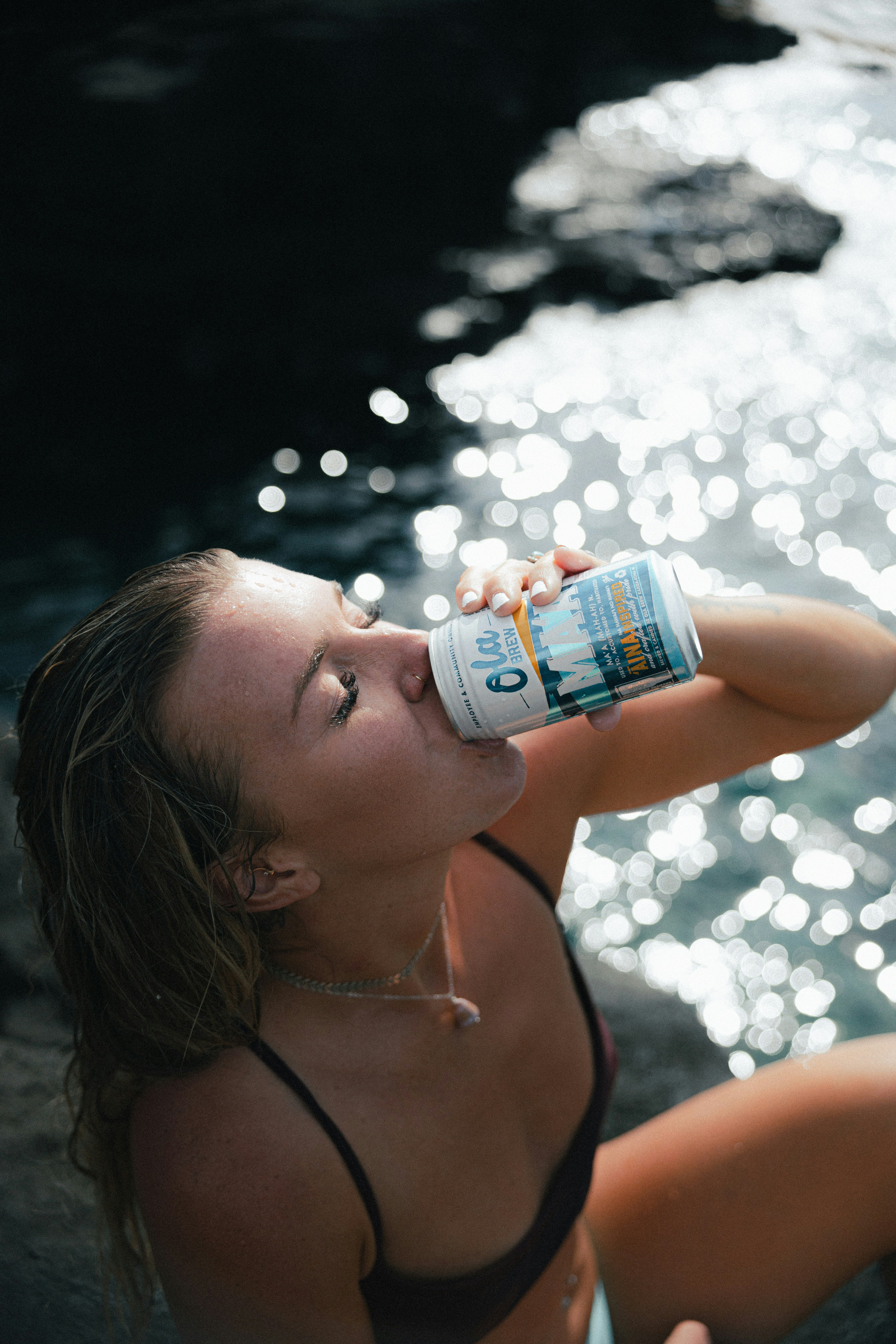 A candid photograph captures a woman in a black bikini taking a drink from a can beside sunlit, glittering water.