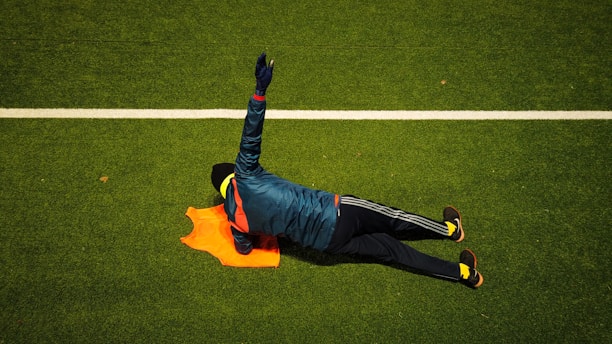 A person wearing a blue jacket and dark pants lies on a grassy sports field. The individual has one arm extended above their head and is near an orange training vest. The field features a white line, suggesting a football or soccer setting.