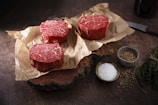 Three raw beef steaks are placed on crumpled parchment paper on a rustic wooden surface. Next to them are small bowls containing salt and peppercorns. A knife and fresh herbs are also visible, arranged neatly to the side.