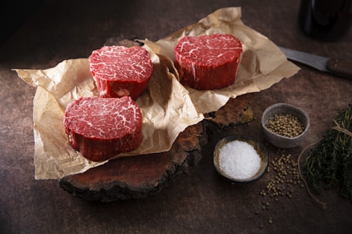 Three raw beef steaks are placed on crumpled parchment paper on a rustic wooden surface. Next to them are small bowls containing salt and peppercorns. A knife and fresh herbs are also visible, arranged neatly to the side.