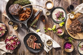 An overhead view of a rustic wooden table filled with an assortment of delicious, hearty foods. There is a cast iron skillet with a large cooked steak and seasoned potato wedges, surrounded by fresh sprigs of rosemary. A black plate nearby holds sliced steak. To the right, there is a wooden board with shredded meat. A plate with a tortilla topped with shredded cabbage, herbs, and a bowl of crumbled cheese. Lime wedges and small bowls of various spices and dips are scattered throughout. A salad with walnuts and radicchio adds a pop of color.