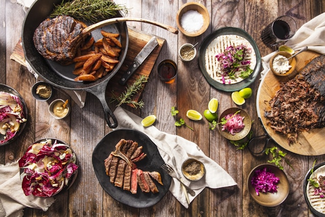 An overhead view of a rustic wooden table filled with an assortment of delicious, hearty foods. There is a cast iron skillet with a large cooked steak and seasoned potato wedges, surrounded by fresh sprigs of rosemary. A black plate nearby holds sliced steak. To the right, there is a wooden board with shredded meat. A plate with a tortilla topped with shredded cabbage, herbs, and a bowl of crumbled cheese. Lime wedges and small bowls of various spices and dips are scattered throughout. A salad with walnuts and radicchio adds a pop of color.
