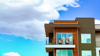 A modern apartment building features a balcony with two round chairs and sleek railings. The facade is a combination of dark green and wood paneling with large windows. The background is a vibrant blue sky with fluffy white clouds.