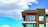 A modern apartment building features a balcony with two round chairs and sleek railings. The facade is a combination of dark green and wood paneling with large windows. The background is a vibrant blue sky with fluffy white clouds.