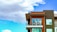 A modern apartment building features a balcony with two round chairs and sleek railings. The facade is a combination of dark green and wood paneling with large windows. The background is a vibrant blue sky with fluffy white clouds.