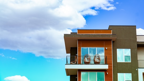 A modern apartment building features a balcony with two round chairs and sleek railings. The facade is a combination of dark green and wood paneling with large windows. The background is a vibrant blue sky with fluffy white clouds.