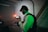 A person wearing a cap operates a professional camera setup indoors. The scene is dimly lit with ambient green lighting. There is a table nearby, and the background includes an industrial setting with a garage door and a staircase.