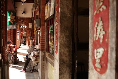 A narrow alleyway with brick walls adorned with red signs featuring calligraphy. Two people, one seated and the other standing, appear engaged in conversation, with a bicycle nearby. The alley is lit by warm, diffused light creating a cozy atmosphere.