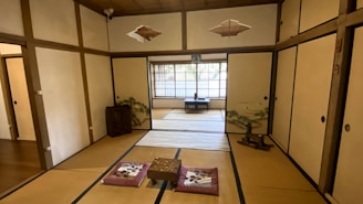 A traditional Japanese room featuring tatami mats on the floor and sliding shoji doors with painted designs. The room includes low seating cushions around a central wooden table. There is a small decorative chest and a potted plant in the corner. Natural light filters in through a large window with a view of bamboo.