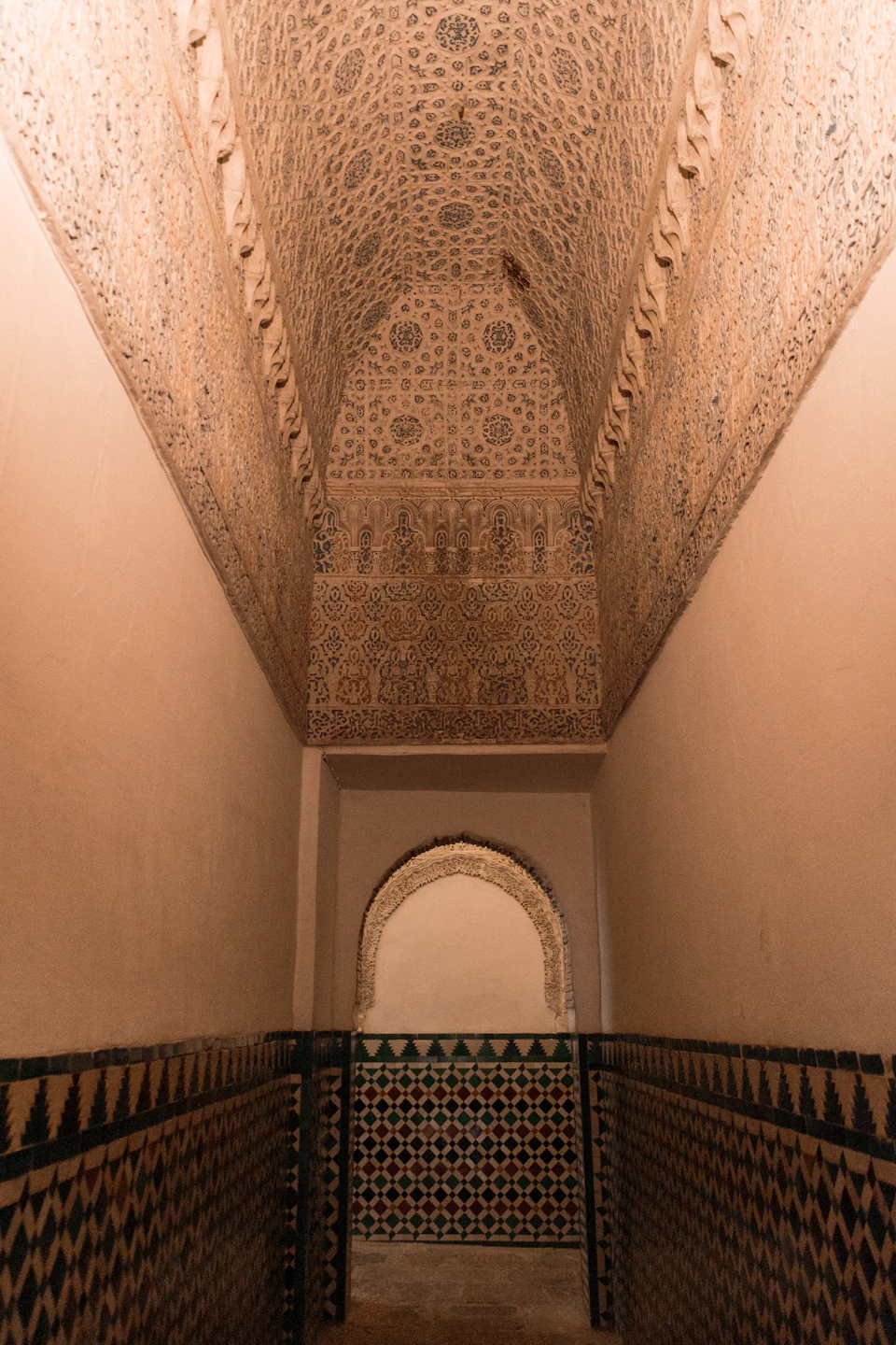 The image shows a narrow hallway with a high ceiling, adorned with intricate patterns and designs. The walls are covered in tiles that display a variety of colors and patterns, creating a visually appealing scene. A large archway is situated at one end of the hallway, adding to its architectural charm. The perspective of the photo is taken from below, looking up towards the ceiling, which enhances the sense of depth and grandeur within the space.