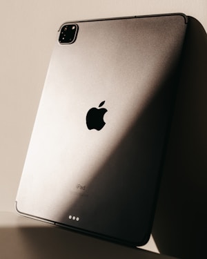 The image features the back of a sleek metallic tablet, prominently displaying the logo of a major technology brand. Soft lighting creates a play of shadows on the surface, enhancing its modern and sophisticated design.