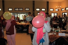 A group of people are in a room with tables covered with tablecloths, bottles, and papers. They are engaged in an activity involving blowing up balloons. Some individuals are wearing hijabs, and everyone seems focused on participating.