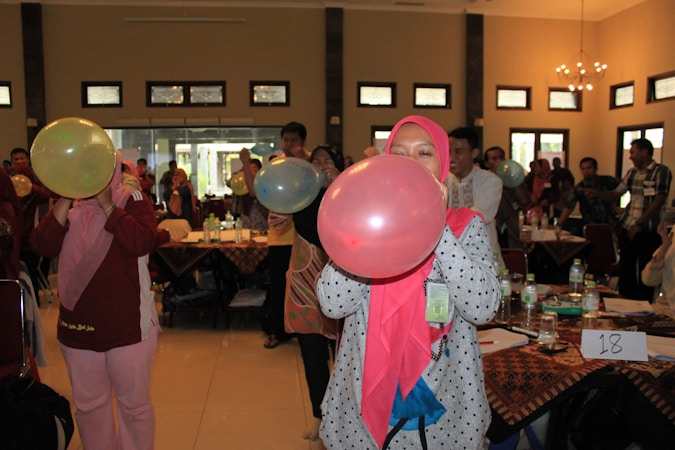 A group of people are in a room with tables covered with tablecloths, bottles, and papers. They are engaged in an activity involving blowing up balloons. Some individuals are wearing hijabs, and everyone seems focused on participating.