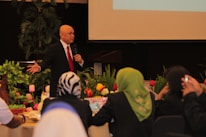 A man wearing a black suit, white shirt and red tie is standing and speaking into a microphone. He is in a room decorated with colorful tulips and green plants. Several people are seated around tables, facing him, and listening attentively.