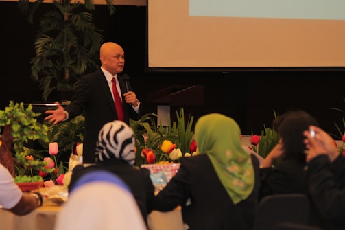 A man wearing a black suit, white shirt and red tie is standing and speaking into a microphone. He is in a room decorated with colorful tulips and green plants. Several people are seated around tables, facing him, and listening attentively.