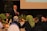 A man wearing a black suit, white shirt and red tie is standing and speaking into a microphone. He is in a room decorated with colorful tulips and green plants. Several people are seated around tables, facing him, and listening attentively.