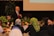 A man wearing a black suit, white shirt and red tie is standing and speaking into a microphone. He is in a room decorated with colorful tulips and green plants. Several people are seated around tables, facing him, and listening attentively.