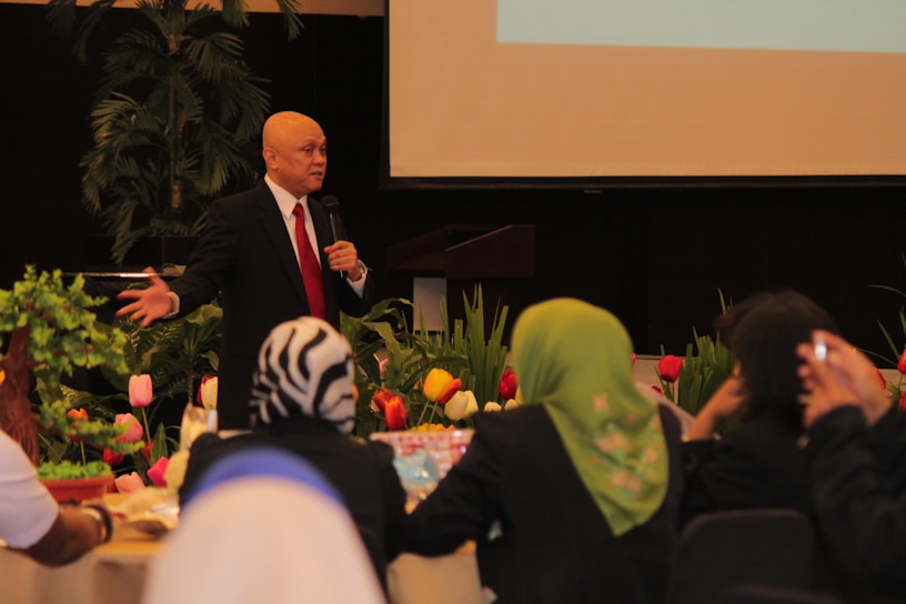 A man wearing a black suit, white shirt and red tie is standing and speaking into a microphone. He is in a room decorated with colorful tulips and green plants. Several people are seated around tables, facing him, and listening attentively.