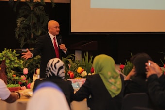 A man wearing a black suit, white shirt and red tie is standing and speaking into a microphone. He is in a room decorated with colorful tulips and green plants. Several people are seated around tables, facing him, and listening attentively.