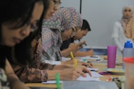 A group of people sitting at a table, focused on writing and taking notes. Several individuals wear patterned hijabs. Various stationery items, such as colorful pens and a pink cup, are scattered on the table. In the background, a woman stands near a whiteboard.