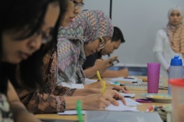 A group of people sitting at a table, focused on writing and taking notes. Several individuals wear patterned hijabs. Various stationery items, such as colorful pens and a pink cup, are scattered on the table. In the background, a woman stands near a whiteboard.