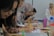 A group of people sitting at a table, focused on writing and taking notes. Several individuals wear patterned hijabs. Various stationery items, such as colorful pens and a pink cup, are scattered on the table. In the background, a woman stands near a whiteboard.