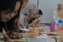 A group of people sitting at a table, focused on writing and taking notes. Several individuals wear patterned hijabs. Various stationery items, such as colorful pens and a pink cup, are scattered on the table. In the background, a woman stands near a whiteboard.