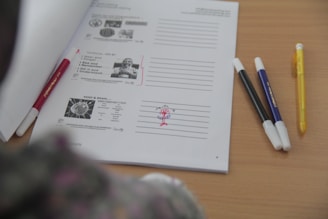 A table with a sheet of paper containing text and diagrams, organized with lined spaces for notes. Several pens of different colors are placed next to the sheet.