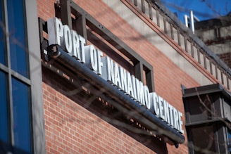 A brick building with a large metal sign displaying 'Port of Nanaimo Centre' in bold white letters. The sign is mounted above a window with blue-tinted glass panels. The sky is clear with a bright blue color, indicating sunny weather.