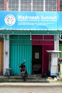 A storefront with a blue and pink metal shutter divided in half, beneath a large blue sign that reads 'Madrosah Sunnah'. The entrance is partially open, revealing an interior space. A parked motorcycle with a visible license plate is positioned in front of the building. To the right, a small structure possibly for shade or storage is present.