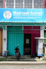 A storefront with a blue and pink metal shutter divided in half, beneath a large blue sign that reads 'Madrosah Sunnah'. The entrance is partially open, revealing an interior space. A parked motorcycle with a visible license plate is positioned in front of the building. To the right, a small structure possibly for shade or storage is present.