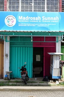 A storefront with a blue and pink metal shutter divided in half, beneath a large blue sign that reads 'Madrosah Sunnah'. The entrance is partially open, revealing an interior space. A parked motorcycle with a visible license plate is positioned in front of the building. To the right, a small structure possibly for shade or storage is present.