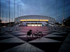 A large, modern building with the illuminated sign 'Jakarta International Velodrome'. The sky is overcast, creating a dim, early evening ambiance. Numerous flag poles surround the area without any flags. In the foreground, a bicycle is parked on the patterned pavement leading up to the entrance.