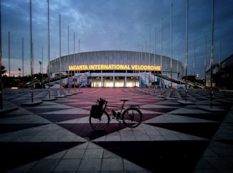 A large, modern building with the illuminated sign 'Jakarta International Velodrome'. The sky is overcast, creating a dim, early evening ambiance. Numerous flag poles surround the area without any flags. In the foreground, a bicycle is parked on the patterned pavement leading up to the entrance.
