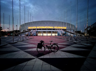 A large, modern building with the illuminated sign 'Jakarta International Velodrome'. The sky is overcast, creating a dim, early evening ambiance. Numerous flag poles surround the area without any flags. In the foreground, a bicycle is parked on the patterned pavement leading up to the entrance.