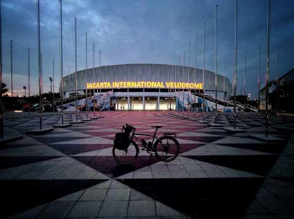 A large, modern building with the illuminated sign 'Jakarta International Velodrome'. The sky is overcast, creating a dim, early evening ambiance. Numerous flag poles surround the area without any flags. In the foreground, a bicycle is parked on the patterned pavement leading up to the entrance.