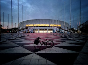 A large, modern building with the illuminated sign 'Jakarta International Velodrome'. The sky is overcast, creating a dim, early evening ambiance. Numerous flag poles surround the area without any flags. In the foreground, a bicycle is parked on the patterned pavement leading up to the entrance.