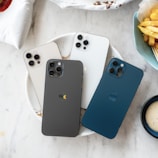 Four smartphones are neatly arranged on a plate, displaying different colors such as white, gray, and blue. The devices have prominent cameras and sit on a marble surface alongside plates and a bowl of fries.