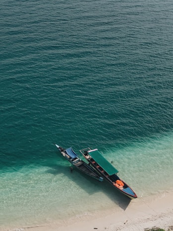 A traditional longtail boat is anchored at the edge of a pristine beach with clear turquoise water gently lapping against the shore. The boat is partially shaded by a green canopy, and orange flotation devices are visible on the deck.