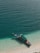 A traditional longtail boat is anchored at the edge of a pristine beach with clear turquoise water gently lapping against the shore. The boat is partially shaded by a green canopy, and orange flotation devices are visible on the deck.