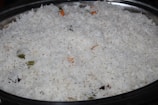 A large serving dish filled with white basmati rice, with a few visible pieces of vegetables such as small green and orange fragments indicating the presence of spices or herbs.
