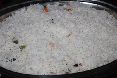 A large serving dish filled with white basmati rice, with a few visible pieces of vegetables such as small green and orange fragments indicating the presence of spices or herbs.