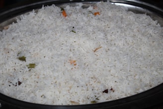 A large serving dish filled with white basmati rice, with a few visible pieces of vegetables such as small green and orange fragments indicating the presence of spices or herbs.