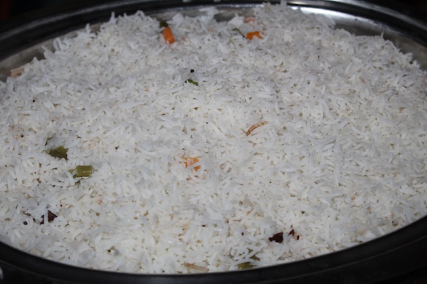 A large serving dish filled with white basmati rice, with a few visible pieces of vegetables such as small green and orange fragments indicating the presence of spices or herbs.