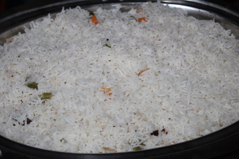 A large serving dish filled with white basmati rice, with a few visible pieces of vegetables such as small green and orange fragments indicating the presence of spices or herbs.